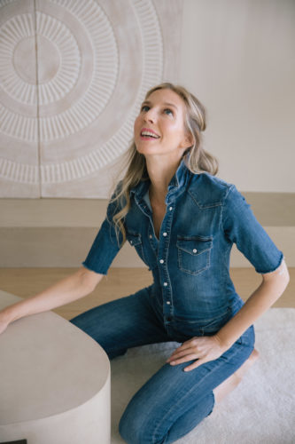 Shelly Lewis in a denim jumpsuit kneels on the floor, smiling and gazing upward. Behind her is a textured, circular wall art. The scene is calm and serene.